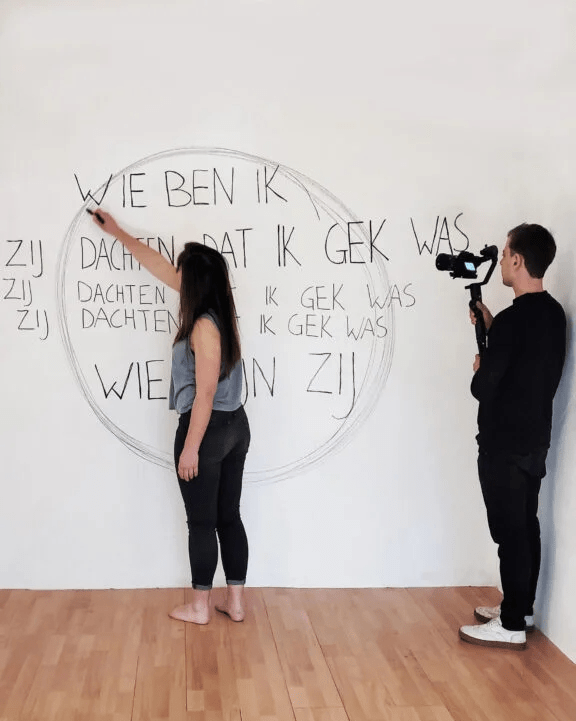 Eva tekent op de muur, ze trekt cirkels over de tekst 'Wie ben ik, Zij dachten dat ik gek was, Wie zijn zij' Daarachter staat Davids Danoss met zijn camera