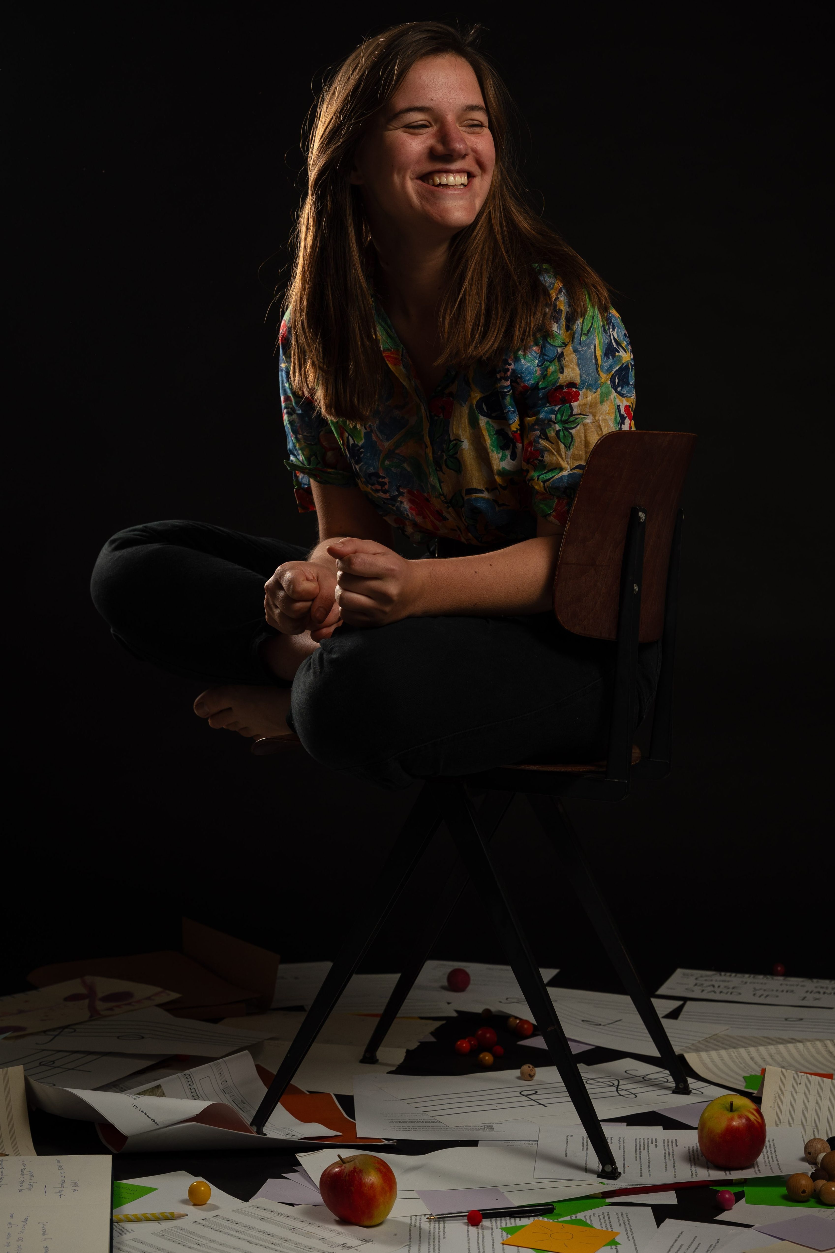 Componist Eva Beunk zit lachend op een stoel de vloer licht vol schetsen van composities, partituren, pennen en appels