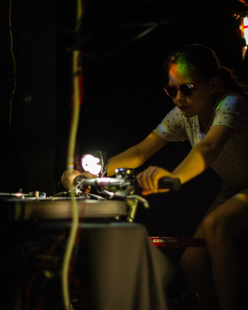 Een meisje is aan het fietsen, de fietslamp schijnt op de platespeler die voor haar staat.