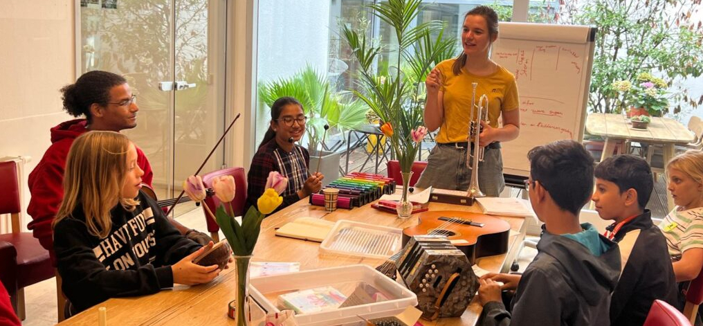 Uitleg bij een compositieworkshop, kinderen zitten aan een tafel vol instrumenten